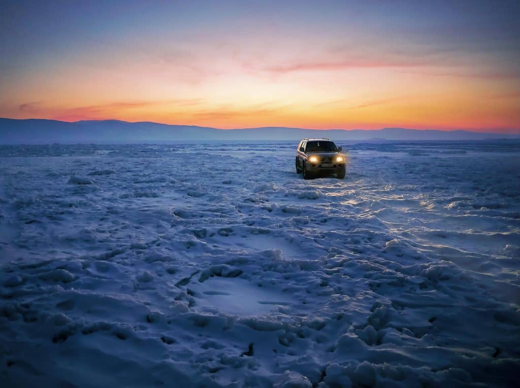 A car with headlights on drives across a vast, snowy, and icy landscape at sunset, with colorful skies and distant mountains in the background.