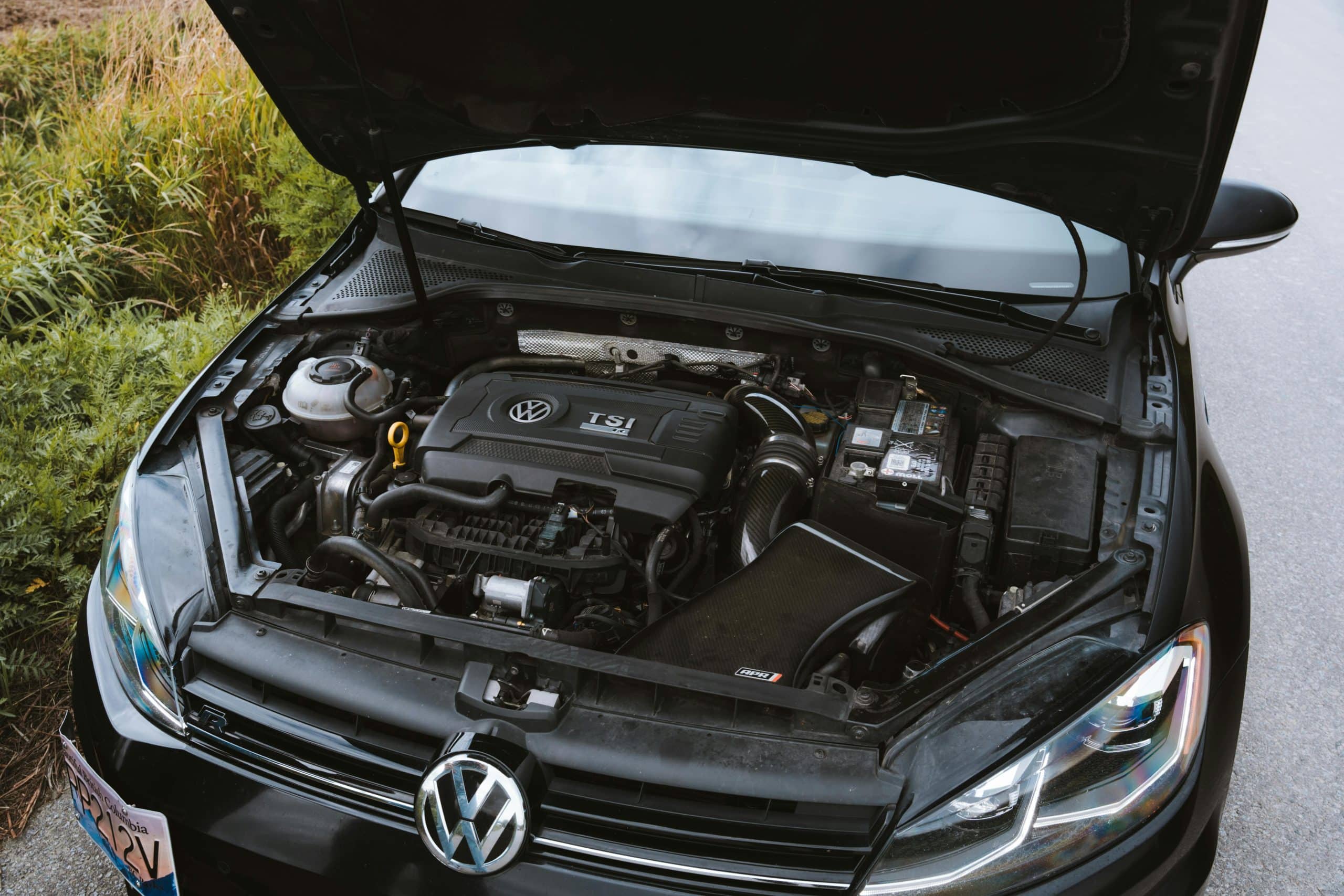 A Volkswagen car with its hood open shows the engine and various engine components, including the TSI engine cover, battery, and fluid reservoirs, parked next to a grassy area.